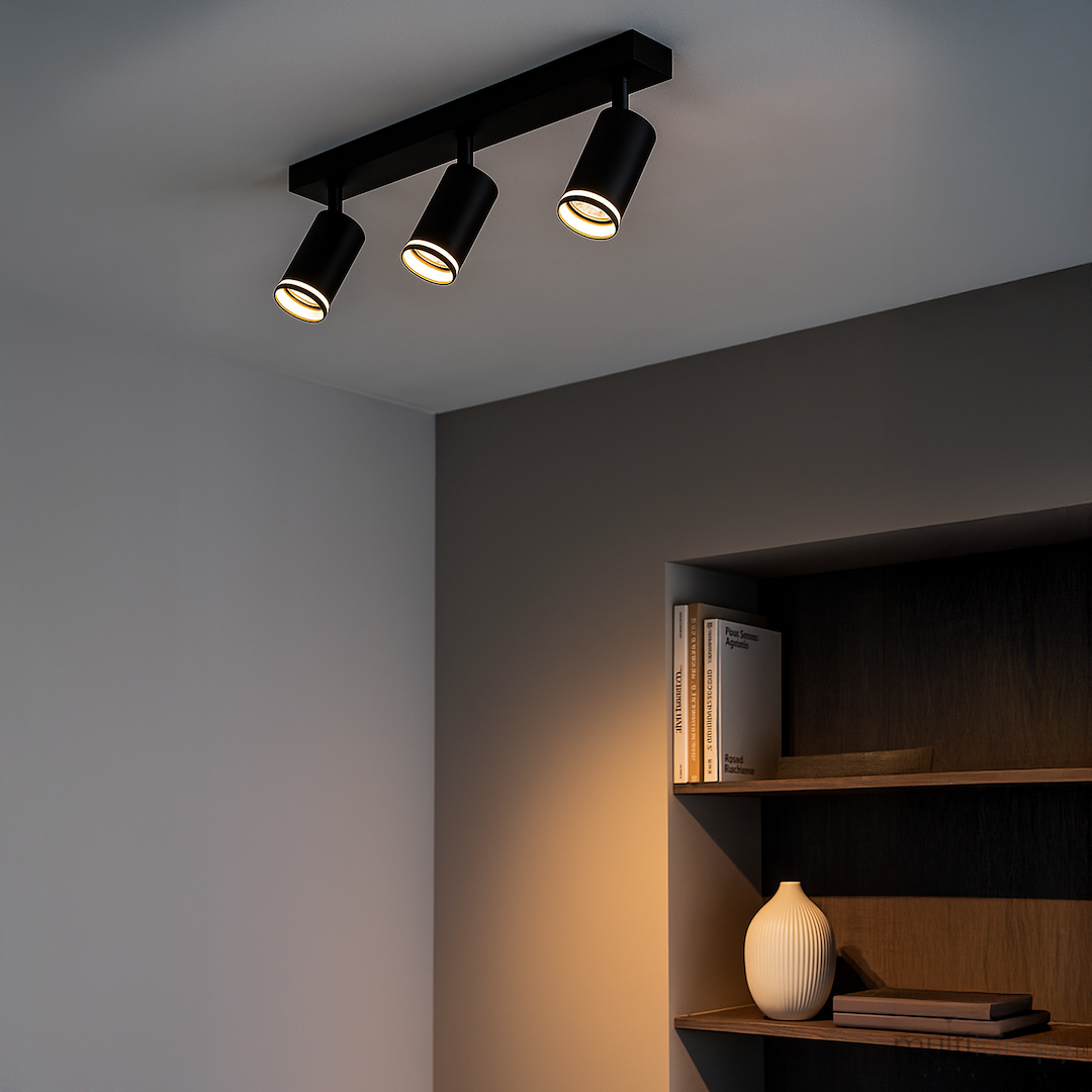Lampa sufitowa Pipe Ring w kolorze czarnym z trzema reflektorami GU10, zamontowana na prostokątnej podstawie. Ujęcie z dołu pokazuje kierunkowe światło, w tle półka z książkami i wazonem na jednolitym, ciemnym tle.
