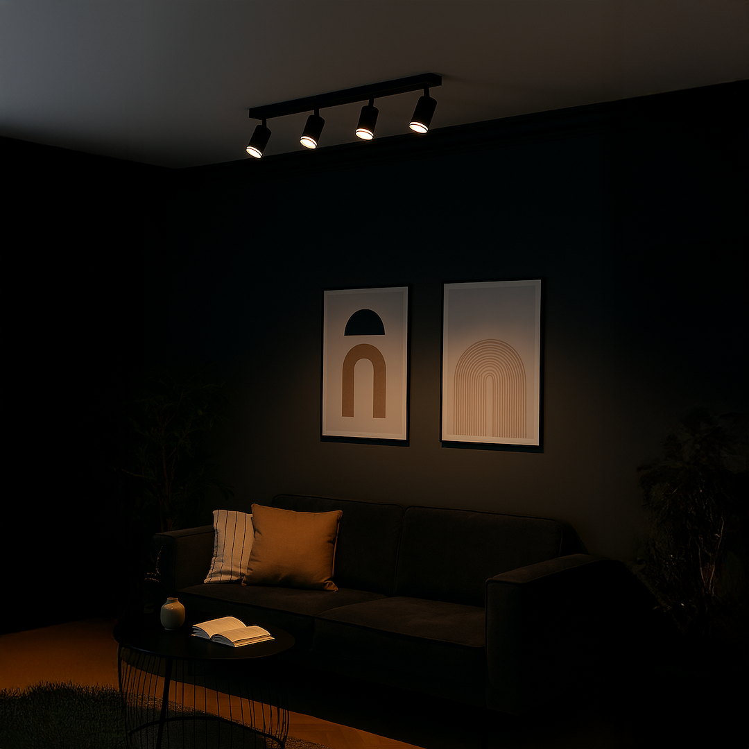 Czarna lampa sufitowa Pipe Ring z czterema reflektorami GU10, montowana na ścianie, o minimalistycznym designie. Ujęcie z boku, w tle widoczne dwa plakaty na ciemnej ścianie oraz sofę z poduszką i stół kawowy z książką.