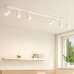 Lampa sufitowa Pipe Ring w kolorze białym, z sześcioma reflektorami na szynie. Reflektory mają cylindryczny kształt i są skierowane w różnych kierunkach. Produkt zamontowany na suficie, w jasnym wnętrzu z półką i oknem w tle.