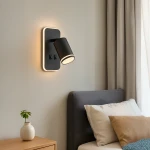 Kinkiet Pipe Ring w kolorze czarnym z pojedynczym źródłem światła GU10 i żarówką LED 6W, zamontowany na ścianie obok łóżka. Posiada okrągły klosz i dwa przyciski na prostokątnej podstawie. W tle widoczna szara pościel i roślina w doniczce.