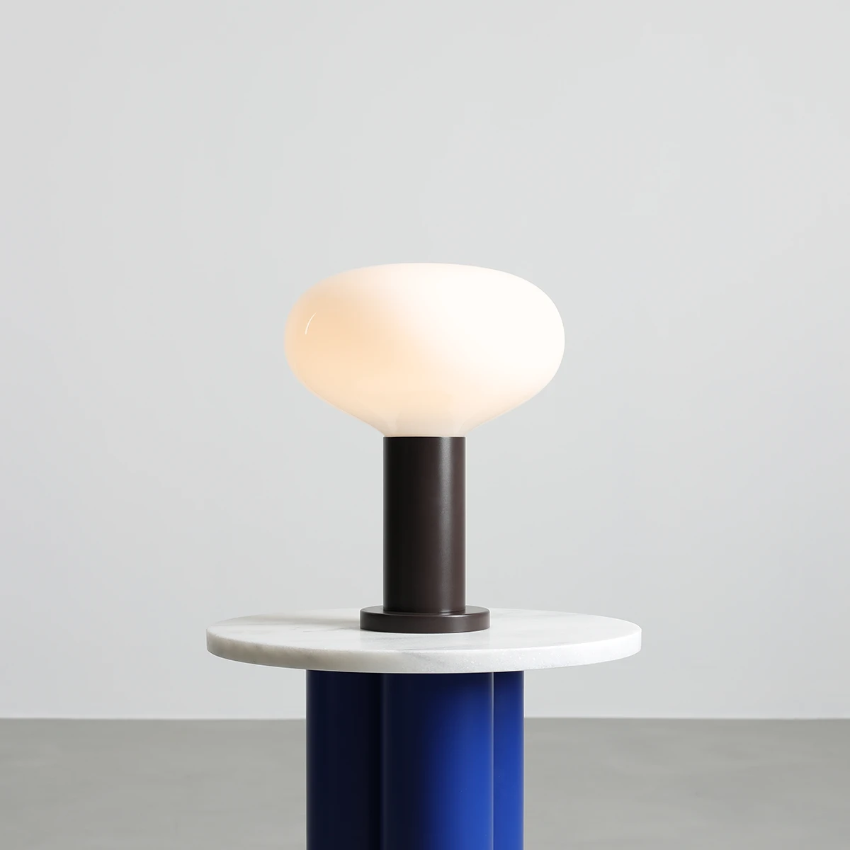 Duża lampa stołowa Flo Mokka z kloszem w kształcie owalnym w kolorze mlecznym, umieszczona na ciemnoszarej podstawie. Lampa stoi na okrągłym, białym blacie, z niebieskim podstawką, na jednolitym tle.
