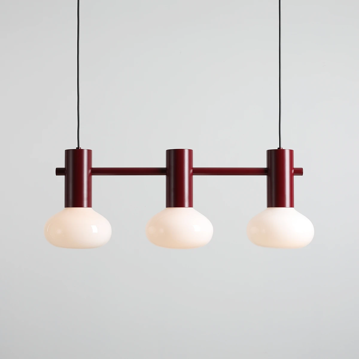 Lampa wisząca Flo Ruby z trzema oprawami E14. Czerwono-brązowa metalowa konstrukcja z okrągłymi, mlecznymi kloszami. Zawieszona na czarnym przewodzie, frontalny widok, minimalistyczny design. Idealna do nowoczesnych wnętrz.