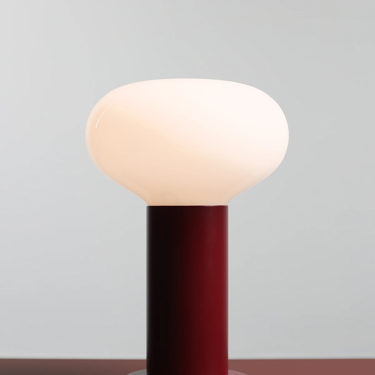 Lampa stołowa Flo Ruby mała z okrągłym kloszem w kolorze białym i cylindryczną podstawą w odcieniu ciemnoczerwonym. Ujęcie frontalne, ukazujące detale kształtu klosza oraz fakturę podstawy.