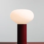 Lampa stołowa Flo Ruby mała z okrągłym kloszem w kolorze białym i cylindryczną podstawą w odcieniu ciemnoczerwonym. Ujęcie frontalne, ukazujące detale kształtu klosza oraz fakturę podstawy.