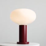Lampa stołowa Flo Ruby z dużą, białą, szklaną kopułą na cylindrycznym, czerwonym korpusie. Ujęcie z boku, stojąca na białym blacie. Idealna do nowoczesnych wnętrz, z miejscem na żarówkę E14.