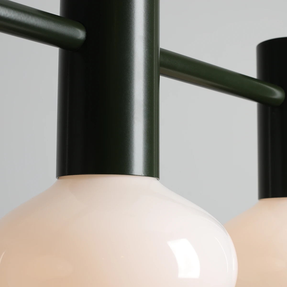 Lampa wisząca Flo Moss z trzema kloszami E14, z metalową, ciemnozieloną konstrukcją i matowym, jasnoróżowym szkłem. Ujęcie z bliska, pokazujące detal mocowania oraz kształt kloszy. Tło jasnoszare, minimalistyczne.