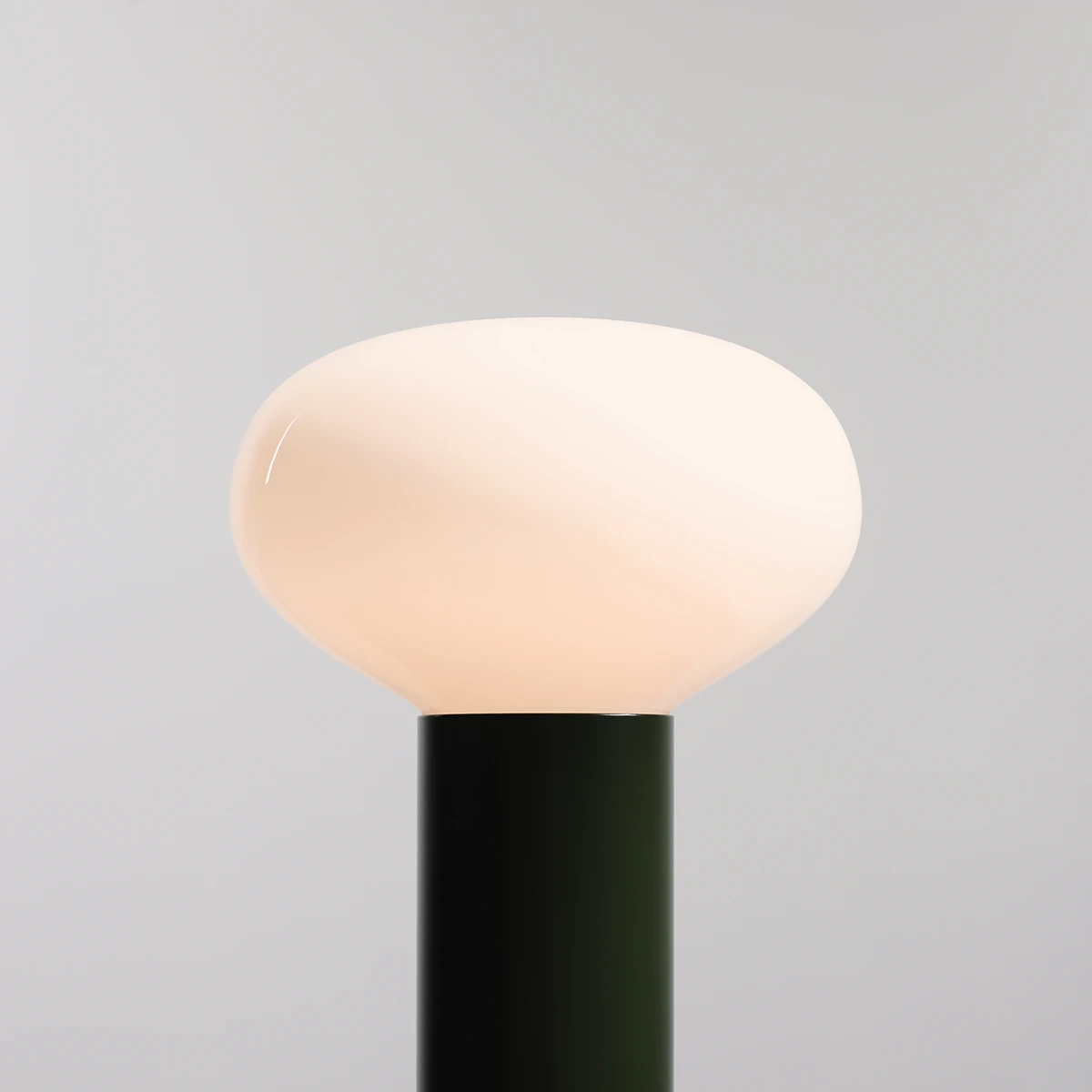 Mała lampa stołowa Flo Moss z białym, zaokrąglonym kloszem z opalowego szkła na czarnym, cylindrycznym stelażu. Produkt ukazany z przodu, z wyraźnym tłem w jasnym kolorze. Wymaga żarówki E14.