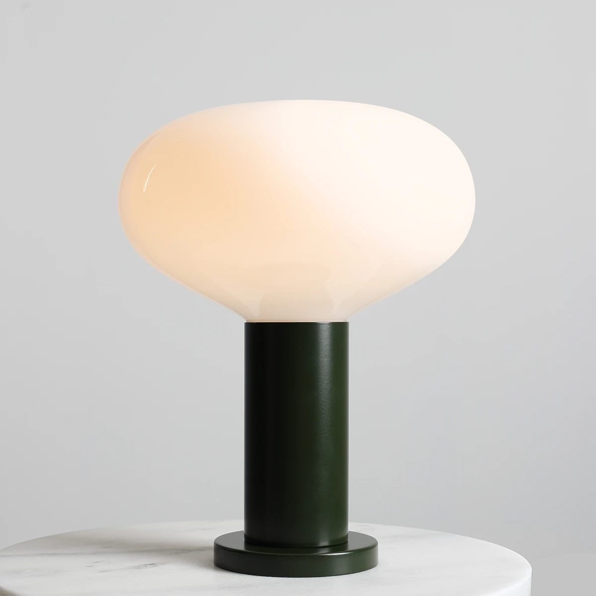 Lampa stołowa Flo Moss w dużym rozmiarze, z okrągłym kloszem w odcieniu mleczno-białym i cylindrycznym, zielonym podstawie. Klosz ma gładką, błyszczącą powierzchnię, a lampa stoi na okrągłej podstawie na białym blacie.