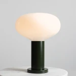 Lampa stołowa Flo Moss w dużym rozmiarze, z okrągłym kloszem w odcieniu mleczno-białym i cylindrycznym, zielonym podstawie. Klosz ma gładką, błyszczącą powierzchnię, a lampa stoi na okrągłej podstawie na białym blacie.