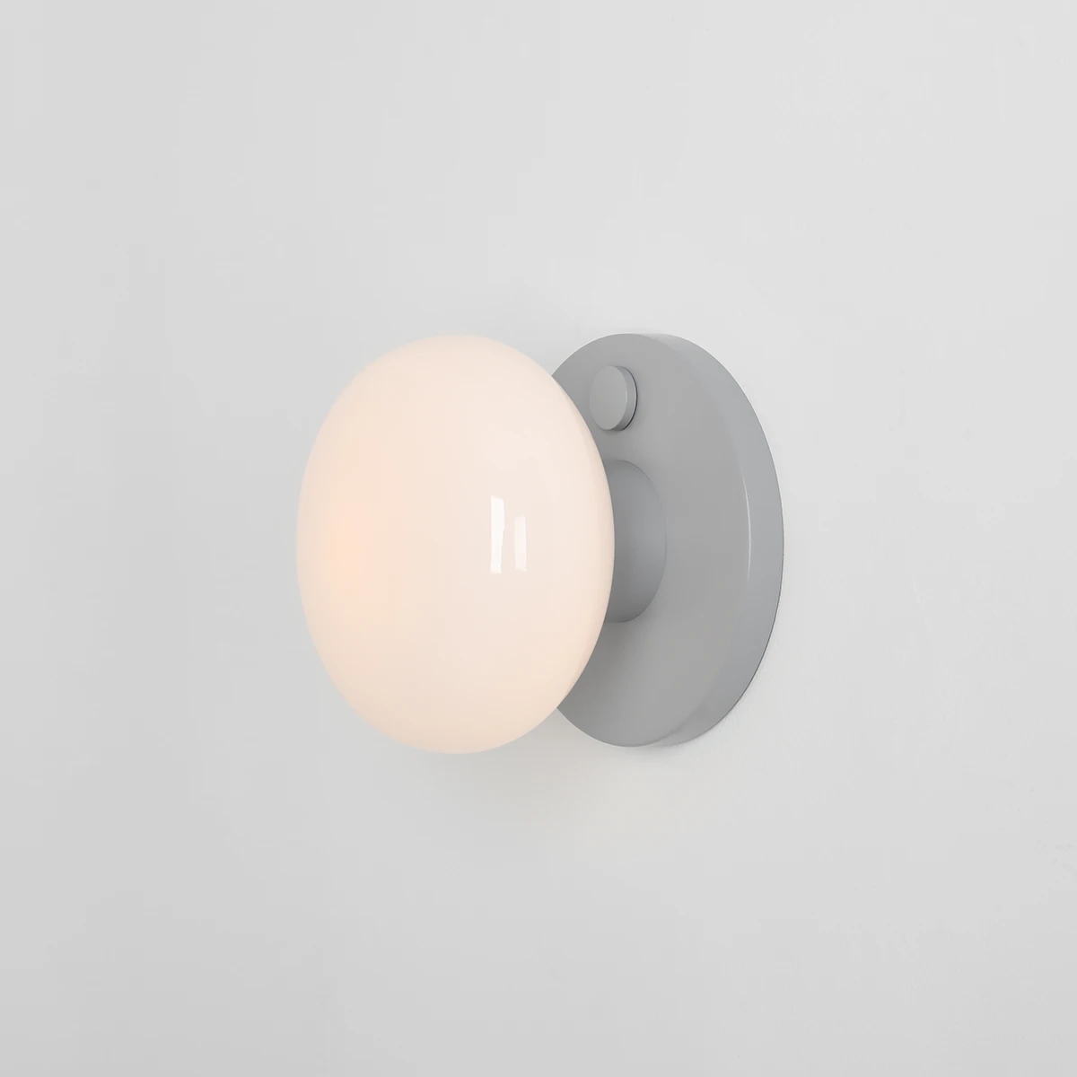 Kinkiet Flo Grey z jedną oprawą E14, z szarym, okrągłym uchwytem i białą, szklaną kulą. Zainstalowany na jasnej ścianie, w pozycji wiszącej, z widocznym włącznikiem na obudowie.