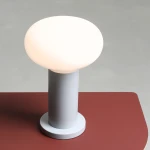 Mała lampa stołowa Flo Grey z kloszem w kształcie kuli w jasnym kolorze, osadzona na cylindrycznej, szarej podstawie. Lampa stoi na czerwonym blacie, z widocznym detalem podstawy. Idealna do nowoczesnych wnętrz.