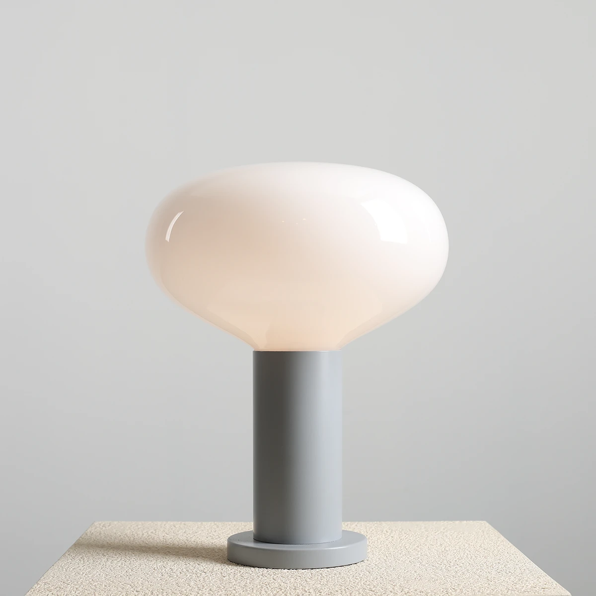 Lampa stołowa Flo Grey z dużą, zaokrągloną kloszem w kolorze mlecznym, umieszczona na szarym, cylindrycznym stelażu. Produkt stoi na jasnej, teksturowanej powierzchni, z jednolitym, szarym tłem. Wymiary klosza i stelaża nie są podane.