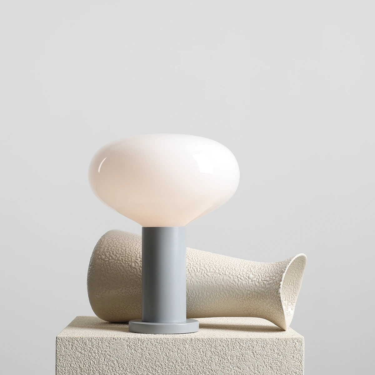 Lampa stołowa Flo Grey, duża, z mlecznobiałym kloszem o opływowym kształcie, zamontowana na szarej podstawie. Tło jasnoszare. Obok widoczny element dekoracyjny w kształcie wazonu.