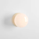 Kinkiet Flo Cream z oprawką E14, o zaokrąglonym, matowym kloszu w kolorze kremowym i dyskretnym, okrągłym mocowaniu. Produkt zamontowany na białej ścianie, stonowane tło podkreśla jego minimalistyczny design.