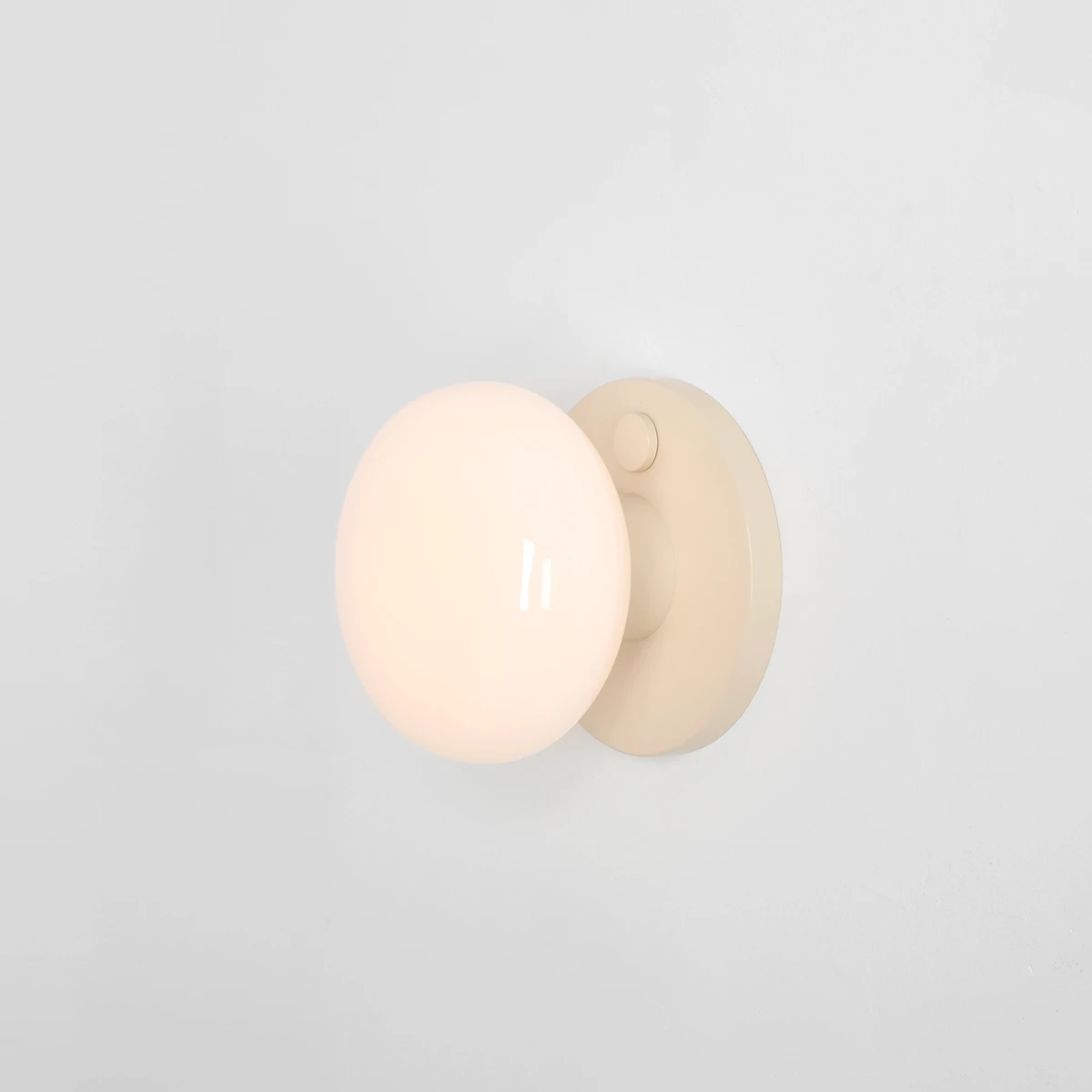 Kinkiet Flo Cream z okrągłą, mleczną kulą na beżowej podstawie, zamontowany na jasnej ścianie. Źródło światła E14, prosty, nowoczesny design. Widoczny przycisk włącznika na podstawie.