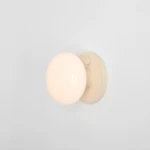 Kinkiet Flo Cream z okrągłą, mleczną kulą na beżowej podstawie, zamontowany na jasnej ścianie. Źródło światła E14, prosty, nowoczesny design. Widoczny przycisk włącznika na podstawie.
