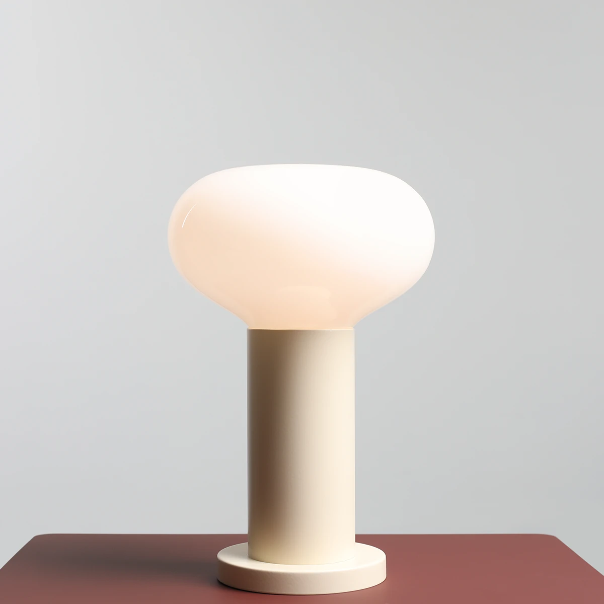 Mała lampa stołowa Flo Cream z kloszem w kształcie owalnym w kolorze mlecznym, osadzona na cylindrycznej, jasnej podstawie. Ujęcie z boku, ukazujące gładką powierzchnię i minimalistyczny design. Tło w odcieniu szaro-beżowym.