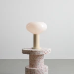 Lampa stołowa Flo Cream z dużą, okrągłą, mlecznobiałą szklaną kloszem na jasnej podstawie. Ustawiona na stołku z teksturowanego materiału w odcieniach bieli i różu. Klosz o gładkiej powierzchni, podstawa cylindryczna.