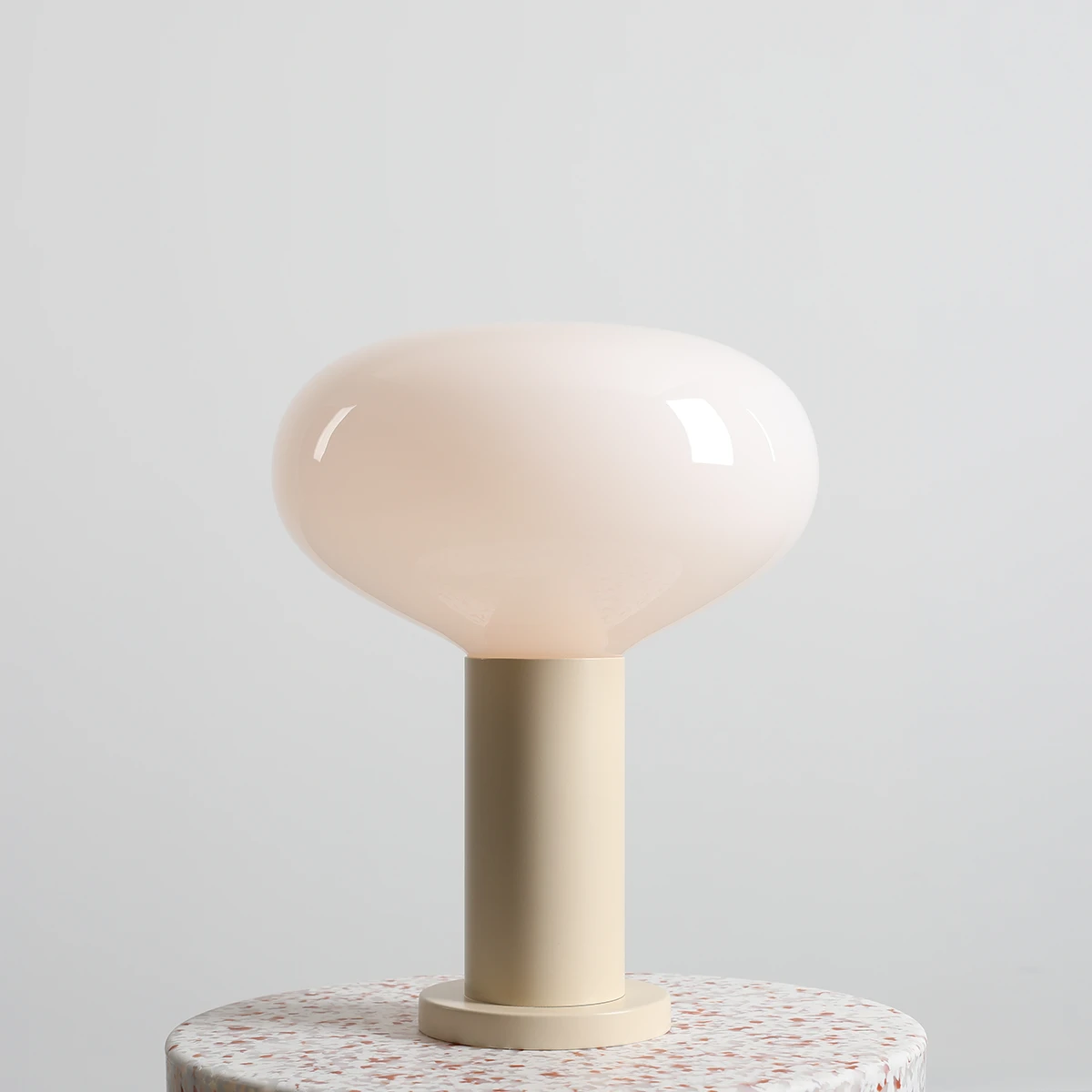 Lampa stołowa Flo Cream z dużą, półprzezroczystą kloszem w kolorze kremowym, umieszczona na jasnej podstawie. Produkt stoi na okrągłym stoliku z dekoracyjnym wzorem. Klosz ma gładką, błyszczącą powierzchnię.
