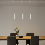 Lampa wisząca Track Pipe w kolorze białym, z trzema cylindrycznymi kloszami, zawieszona na białej szynie. Produkt umieszczony nad drewnianym stołem, w tle widoczne krzesła i wazon z kwiatami.