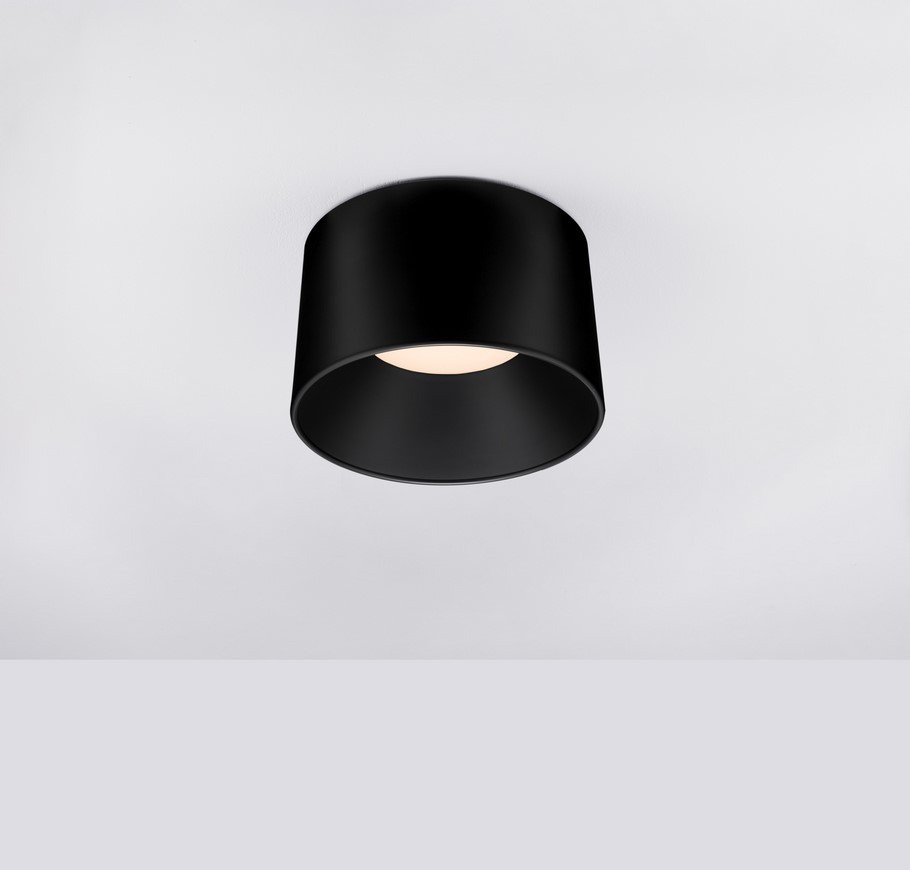 Bajo lampa sufitowa LED 24W w kolorze czarnym, okrągła, zamontowana na suficie. Posiada matowe wykończenie i emitującą światło oprawę w centralnej części. Tło jasne, jednolite.