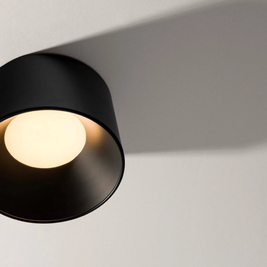 Czarna lampa sufitowa LED Bajo o mocy 24W z okrągłą, białą żarówką. Ujęcie z góry, pokazujące lampę zamontowaną na ścianie, rzucającą cień na gładką powierzchnię. Minimalistyczny design, matowe wykończenie.
