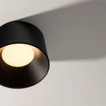 Czarna lampa sufitowa LED Bajo o mocy 24W z okrągłą, białą żarówką. Ujęcie z góry, pokazujące lampę zamontowaną na ścianie, rzucającą cień na gładką powierzchnię. Minimalistyczny design, matowe wykończenie.