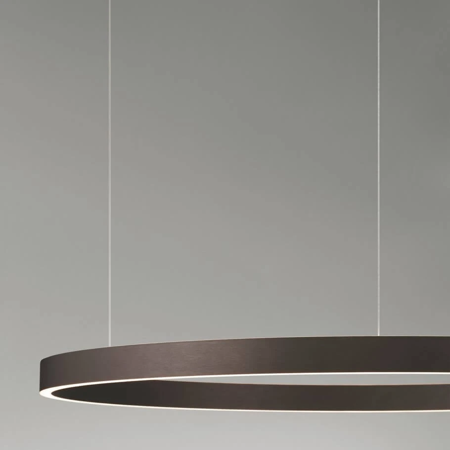 Brązowa lampa wisząca Bari D98 z jedną diodą LED 106W, w kształcie minimalistycznego okręgu, zawieszona na cienkich linkach. Oświetlenie emituje delikatne światło przez wąski pasek na krawędzi. Tło jasnoszare.