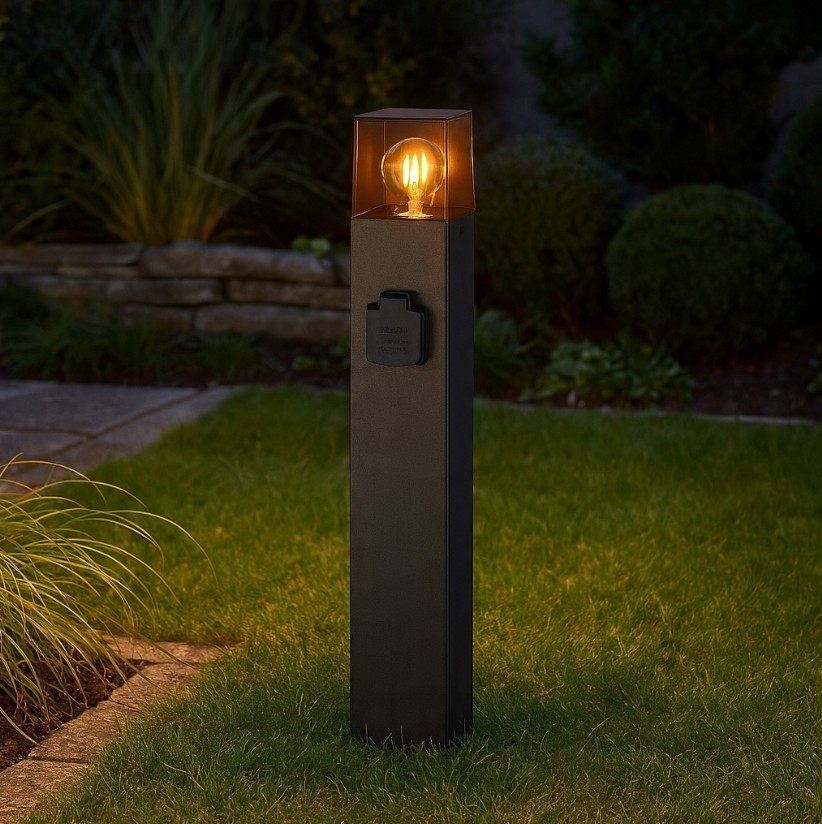 Lampa stojąca ogrodowa Denver H60 w kolorze czarnym, prosta forma prostokątna z przezroczystym kloszem, wbudowanym gniazdem E27. Ujęcie z boku, lampka świeci ciepłym światłem, otoczenie z trawnikiem i krzewami.