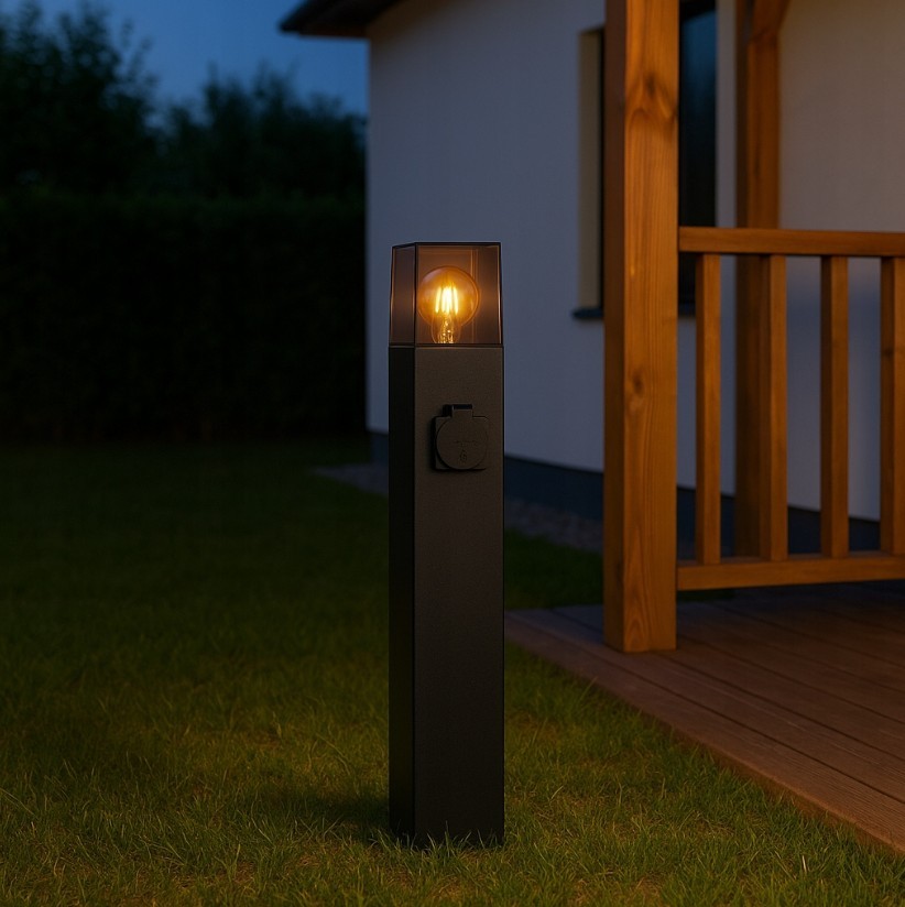 Lampa stojąca ogrodowa Denver H60 w kolorze czarnym, z prostokątną podstawą i przezroczystym kloszem, z widocznym gniazdem E27. Ujęcie z boku, na trawie, z podświetloną żarówką, w tle drewniana weranda i elewacja budynku.