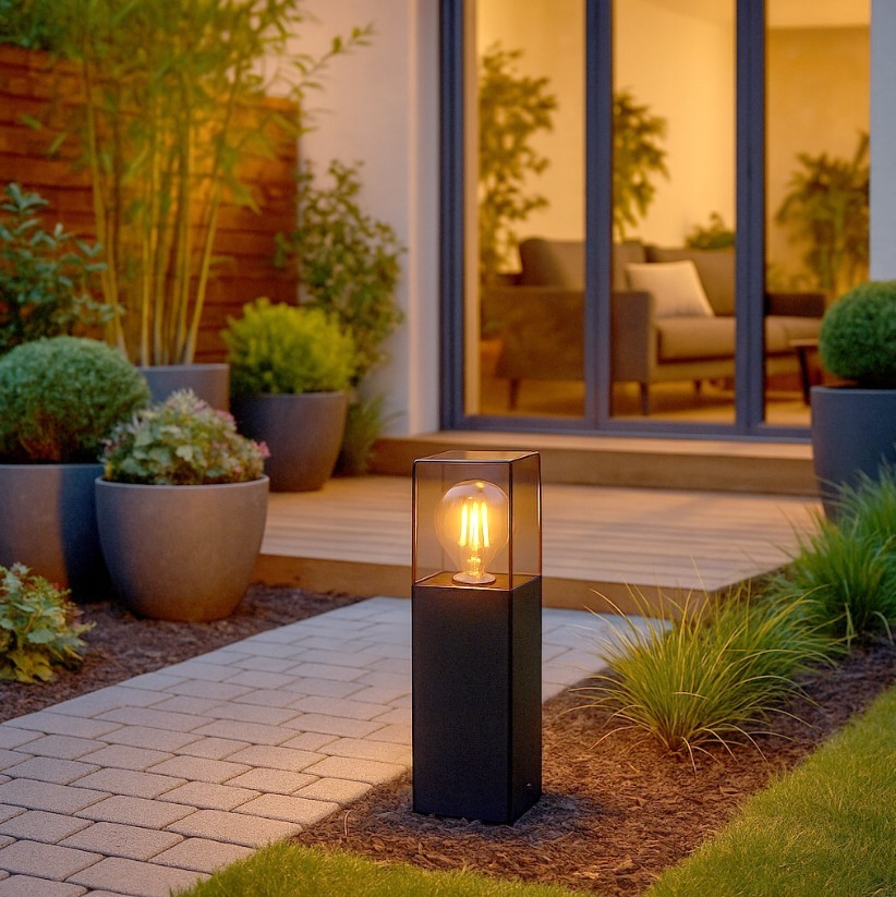 Lampa stojąca ogrodowa Denver H30 w kolorze czarnym z oprawą E27, znajdująca się na ścieżce w ogrodzie. Z przezroczystą osłoną, podświetlona ciepłym światłem, otoczona roślinnością i donicami, z drewnianym tarasem w tle.