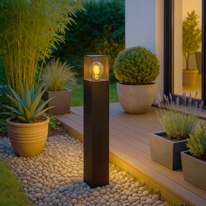 Lampa stojąca ogrodowa Denver H60 w kolorze czarnym, z prostokątną podstawą i przezroczystą osłoną na żarówkę E27, umieszczona na drewnianym tarasie, otoczona donicami z roślinami, na tle zieleni.