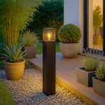 Lampa stojąca ogrodowa Denver H60 w kolorze czarnym, z prostokątną podstawą i przezroczystą osłoną na żarówkę E27, umieszczona na drewnianym tarasie, otoczona donicami z roślinami, na tle zieleni.