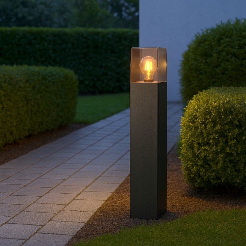 Lampa stojąca ogrodowa Denver H60 w kolorze czarnym, z prostokątną podstawą i przezroczystym kloszem. Widoczna żarówka E27 emitująca ciepłe światło, umieszczona w górnej części. Ustawiona na ścieżce w otoczeniu trawnika i żywopłotu.