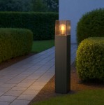 Lampa stojąca ogrodowa Denver H60 w kolorze czarnym, z prostokątną podstawą i przezroczystym kloszem. Widoczna żarówka E27 emitująca ciepłe światło, umieszczona w górnej części. Ustawiona na ścieżce w otoczeniu trawnika i żywopłotu.