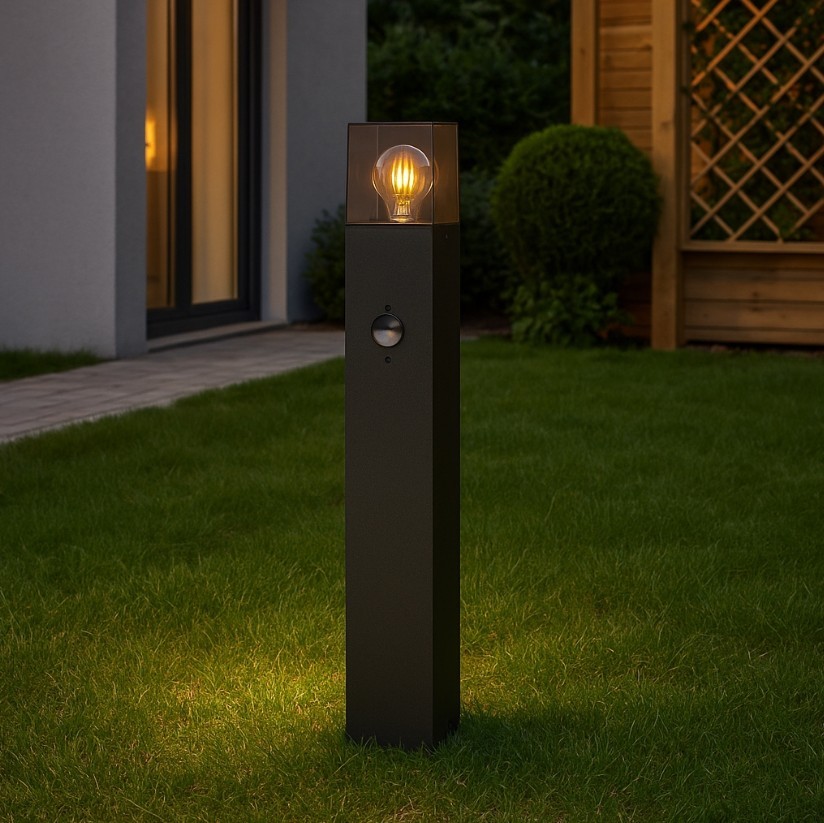 Lampa stojąca ogrodowa Denver H60 w kolorze czarnym, z prostokątnym korpusem i przezroczystą osłoną na żarówkę E27. Widoczny czujnik ruchu na froncie, ustawiona na trawniku, w tle widać budynek i ogrodzenie.