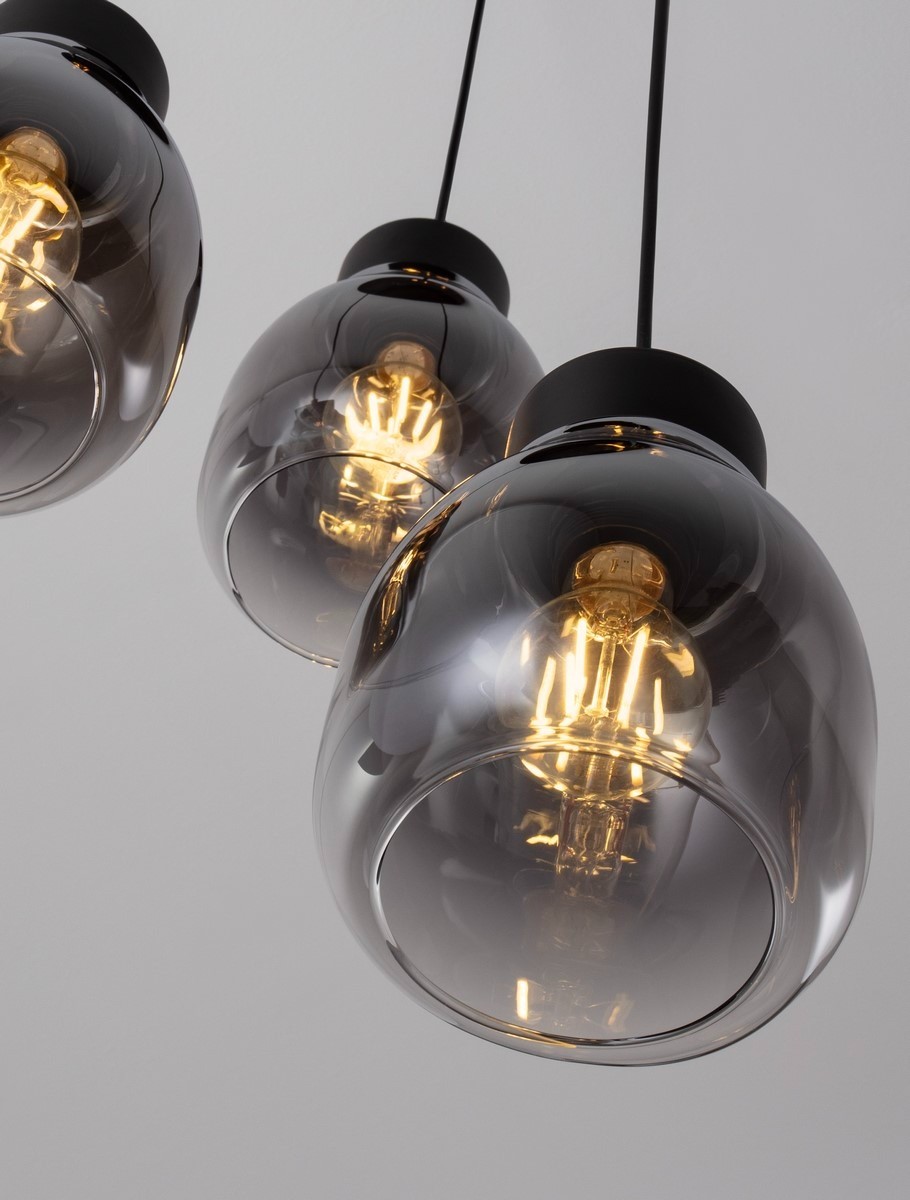 Lampa wisząca Moche w kolorze czarnym z trzema kloszami w kształcie owalnym, wykonana z przezroczystego szkła o ciemnym odcieniu, zawieszona na cienkich linkach. Widoczne żarówki E27 w środku, emitujące ciepłe światło.