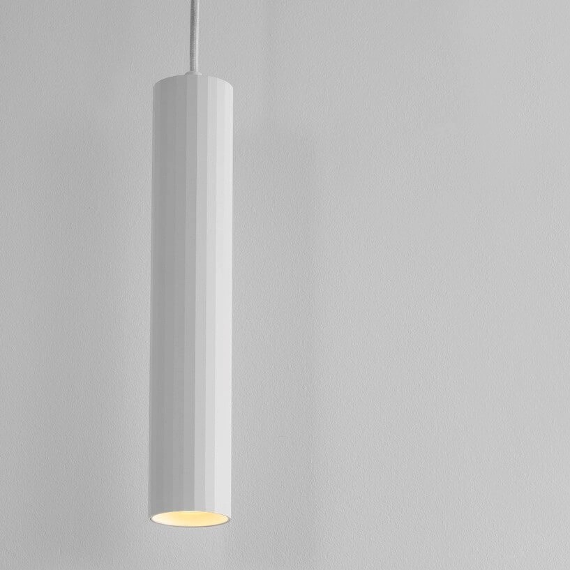 Lampa wisząca Nazca w białym kolorze, cylindryczny kształt z teksturowaną powierzchnią. Umieszczona w pionie, z widocznym źródłem światła na dole. Tło w odcieniu szarości, minimalistyczne i neutralne. Idealna do nowoczesnych wnętrz.