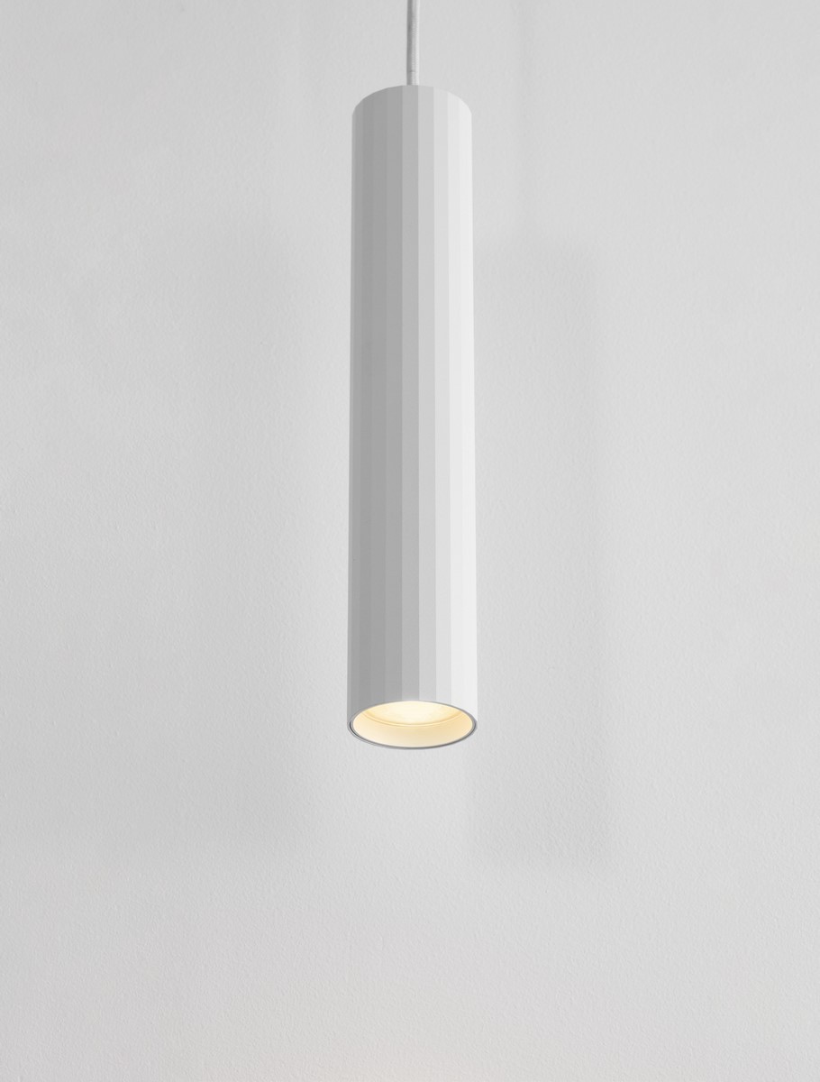 Lampa wisząca Nazca w białym kolorze, cylindryczna, z fakturą w pionowe żłobienia. Umieszczona na białym tle, z widoczną oprawą na żarówkę GU10. Kąt ujęcia z dołu, ukazujący górną część lampy i źródło światła.