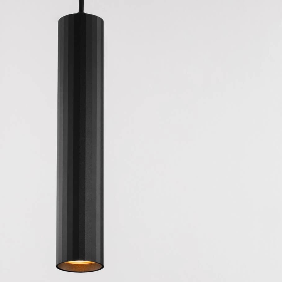 Czarna lampa wisząca Nazca z jedną oprawą GU10, o cylindrycznym kształcie z pionowymi rowkami. Wnętrze lampy w kolorze mosiądzu, zwisająca z cienkiego kabla, umieszczona na jasnym tle. Idealna do nowoczesnych aranżacji wnętrz.