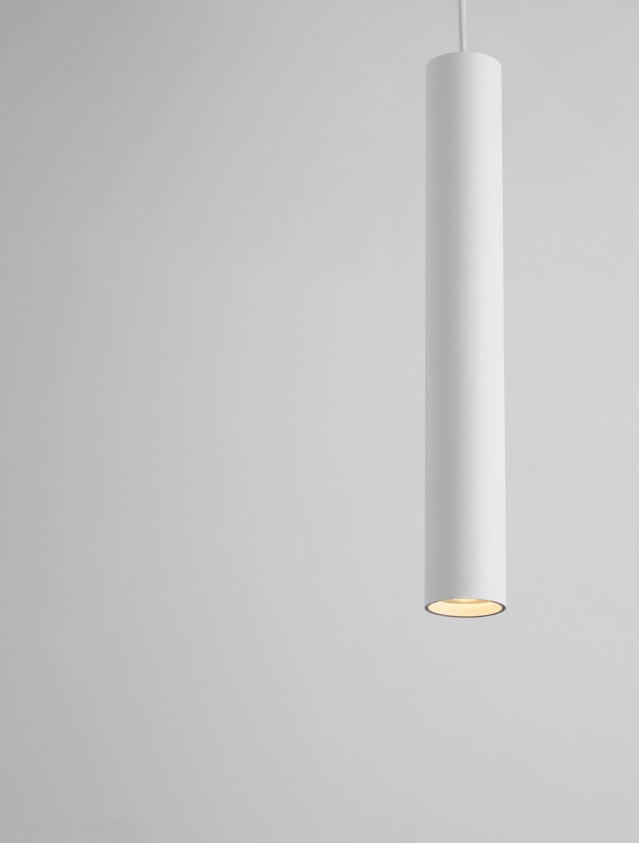 Lampa wisząca Nazca w kolorze białym, o cylindrycznym kształcie. Zawieszona na cienkim przewodzie, z widocznym źródłem światła GU10 u dołu. Tło jasne, minimalistyczne, podkreślające prosty design lampy.