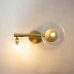 Kinkiet Avola 2xG9 w złotym kolorze z transparentnymi kloszami. Ułożony poziomo, z okrągłymi kloszami, jeden z nich oświetlony. Metalowa konstrukcja z okrągłą podstawą montażową. Tło neutralne, jasne.