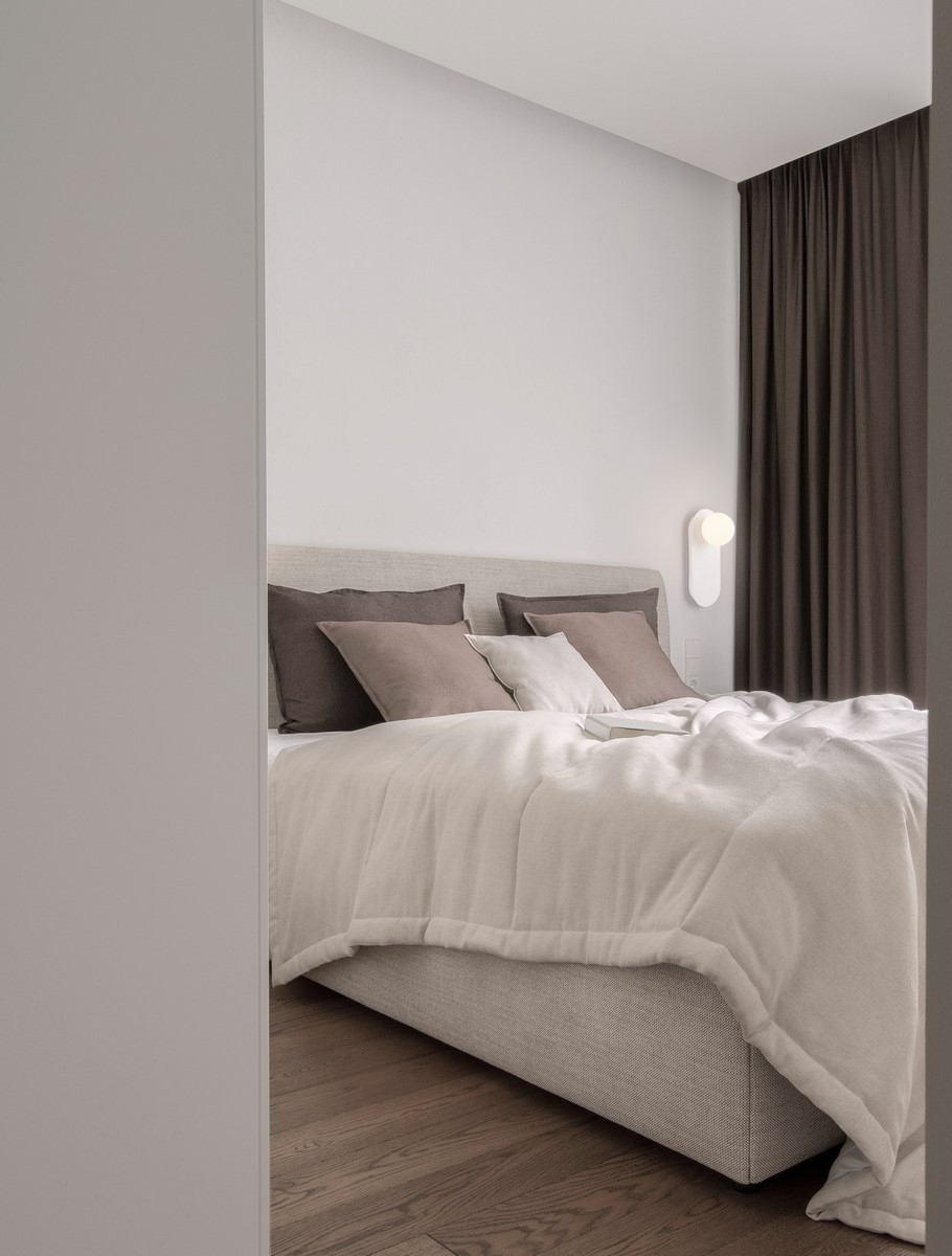 Biały kinkiet Altos 1xG9 zamontowany na ścianie obok łóżka w sypialni. Kinkiet ma minimalistyczny design z okrągłą lampą i prostokątną podstawą. W tle widoczne ciemne zasłony oraz posłanie z jasną narzutą i poduszkami w odcieniach brązu.