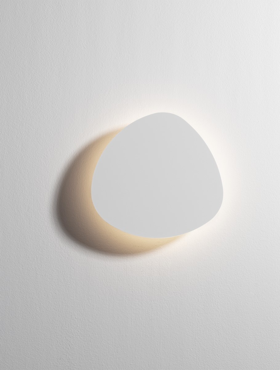 Kinkiet Apure 1xLED 6W w kolorze białym, o asymetrycznym, zaokrąglonym kształcie. Zainstalowany na ścianie, emituje ciepłe światło otaczające. Prosty design idealny do nowoczesnych wnętrz. Tło w jednolitym białym kolorze.