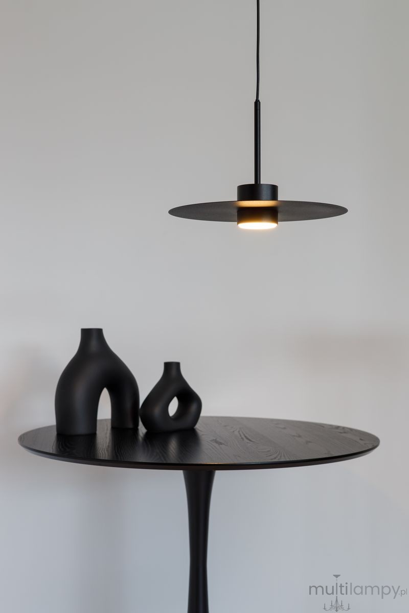 Lampa wisząca Charlie M w kolorze czarnym z jednym źródłem światła GX53, zawieszona na cienkim przewodzie. W tle widoczny okrągły stół z ciemnego drewna oraz trzy ceramiczne wazony o różnych kształtach, również w odcieniach czerni.