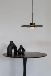 Lampa wisząca Charlie M w kolorze czarnym z jednym źródłem światła GX53, zawieszona na cienkim przewodzie. W tle widoczny okrągły stół z ciemnego drewna oraz trzy ceramiczne wazony o różnych kształtach, również w odcieniach czerni.