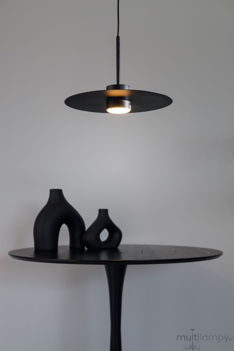 Lampa wisząca Charlie M w kolorze czarnym z oprawą na żarówkę GX53. Posiada okrągły klosz, zawieszona na cienkim przewodzie nad czarnym okrągłym stolikiem, na którym znajdują się trzy czarne wazony o różnych kształtach. Tło w jasnym kolorze.