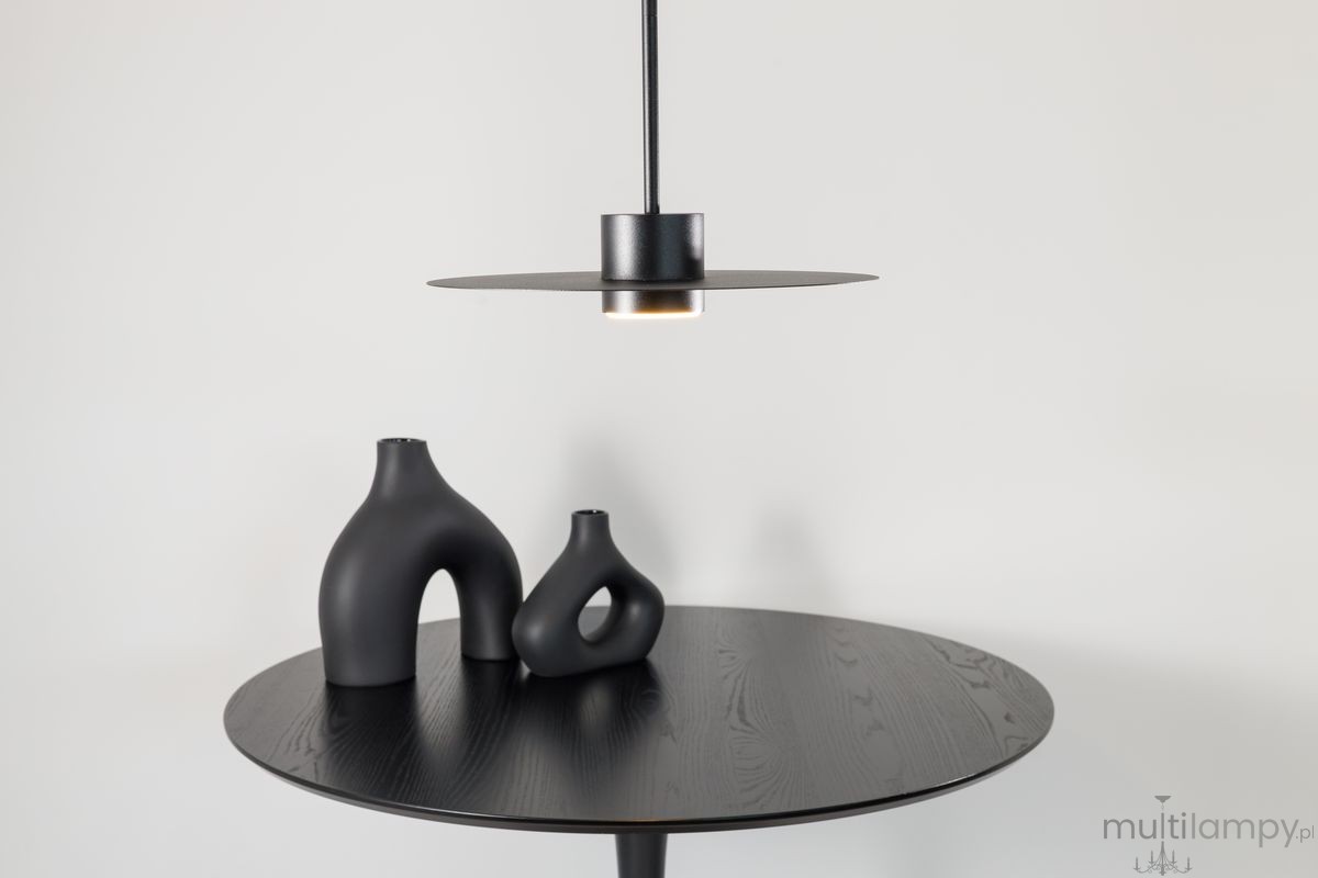 Lampa wisząca Charlie M w kolorze czarnym, z kloszem w formie płaskiego talerza i oprawką na żarówkę GX53. Zawieszona nad okrągłym stołem z ciemnego drewna, obok ceramiczne wazony o organicznych kształtach. Tło jasne, jednolite.