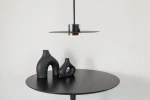 Lampa wisząca Charlie M w kolorze czarnym, z kloszem w formie płaskiego talerza i oprawką na żarówkę GX53. Zawieszona nad okrągłym stołem z ciemnego drewna, obok ceramiczne wazony o organicznych kształtach. Tło jasne, jednolite.