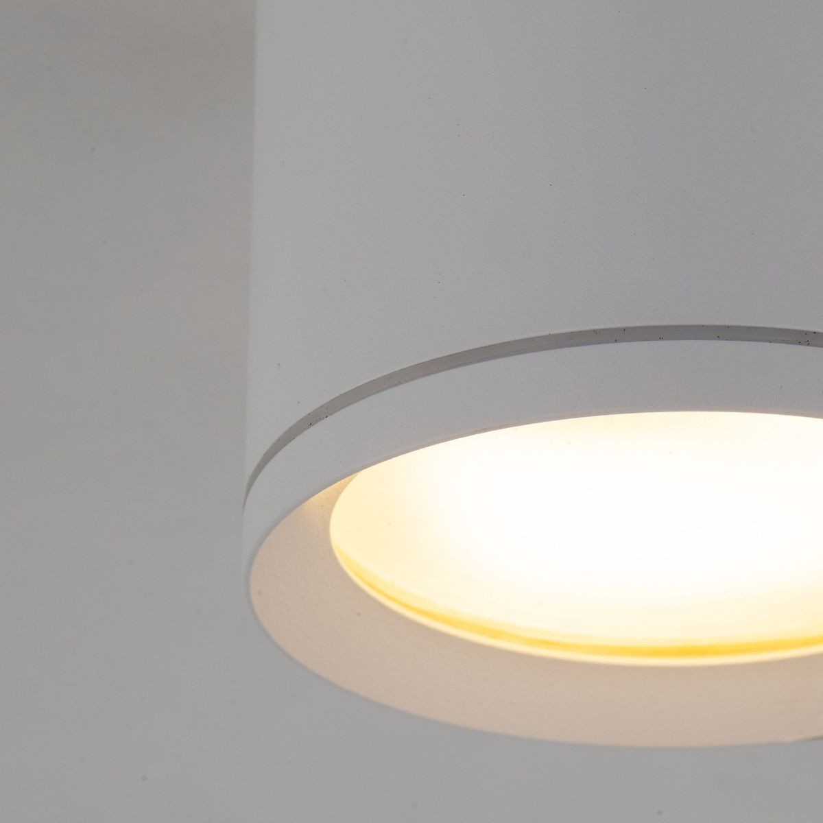 Lampa sufitowa Vivo w kolorze białym z oprawą na żarówkę GX53. Ujęcie z bliska, ukazujące okrągły klosz z matowego materiału oraz emitowane ciepłe światło. Produkt ma kształt cylindryczny, montowany na suficie, z klasą szczelności IP44.